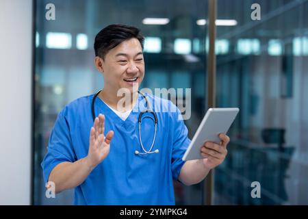 Asian man doctor smiling while using tablet for virtual healthcare consultation. Wearing blue scrubs with stethoscope, communicating effectively, creating positive connection. Stock Photo