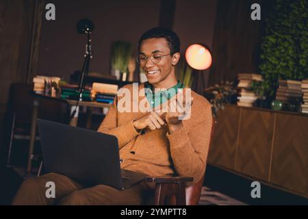 Young man in casual cardigan studying on laptop at home in evening ambient setting Stock Photo