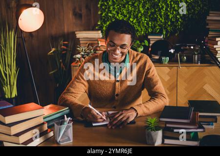 Young student studying at home surrounded by greenery and warm lighting, wearing a casual cardigan, focuses on homework in the evening Stock Photo