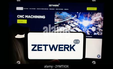 In this photo illustration, a person is holding a cellphone with the logo of Indian company Zetwerk Manufacturing Businesses Pvt. Ltd. in front of webpage. Stock Photo