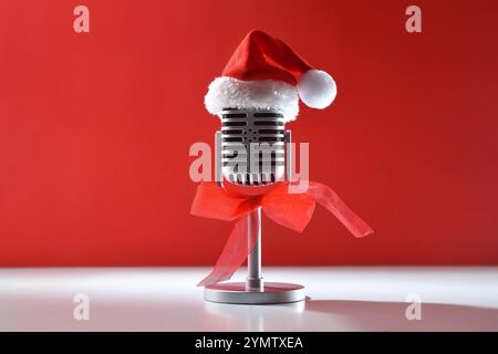Retro microphone for communication decorated with Christmas hat and red bow on white table isolated on red background. Front view. Stock Photo
