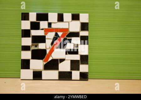 number 7 mosaic house number made from black ,white and red ceramic tiles leaning against a green ceramic tile after being made by hand. Stock Photo