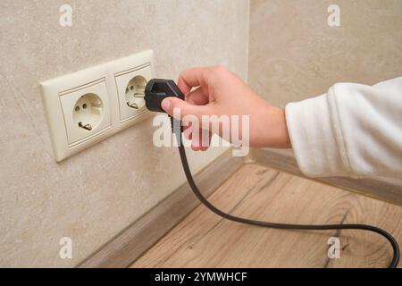 Hand plugging a black electrical plug into a white wall socket. Close-up photography of an electrical connection concept. Stock Photo