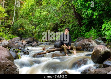 Man sitting on the river while looking around. Young man sitting alone in the middle of rain forest Stock Photo