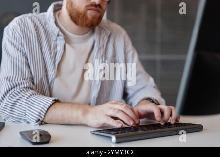 Man working at desk using a keyboard and mouse with office background visible. Focus is on hands typing, suggesting detailed concentration on his task Stock Photo