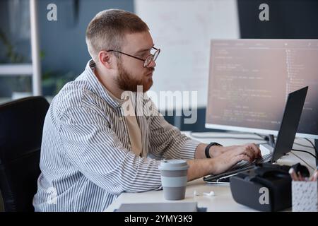 Man with beard intensely focused on coding at desk in modern office space, surrounded by technology and screens Stock Photo