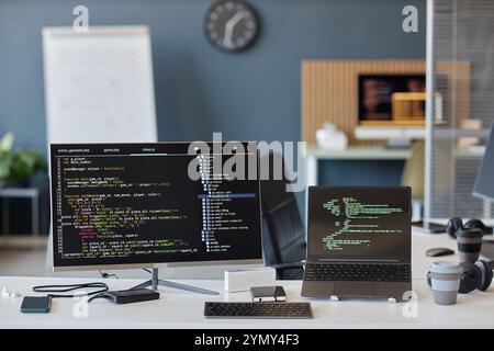 A modern office setup displaying desktop and laptop screens showing code. Background includes a clock and whiteboard, indicating a collaborative work Stock Photo