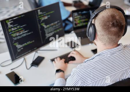 Man wearing headphones sitting at desk working on coding project on dual monitors. Image shows work environment with various devices and tools surroun Stock Photo
