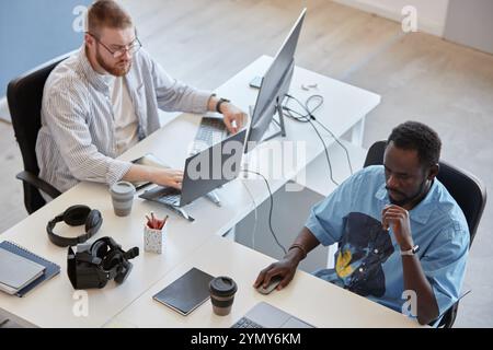 Two colleagues working on computers in a modern office. Both are focused on their tasks, with various gadgets and office supplies on their desks Stock Photo
