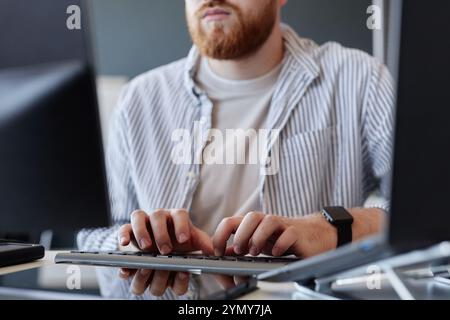 Focused male programmer working on coding projects on dual monitors in contemporary office setting, wearing striped shirt and remaining attentive to t Stock Photo