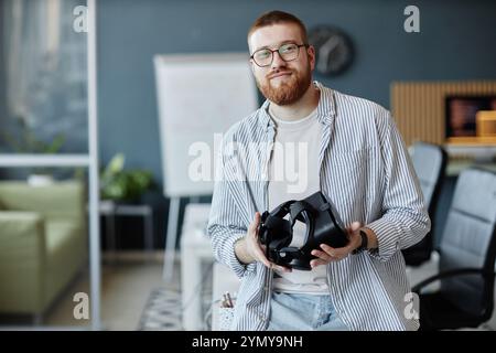 Portrait of bearded man wearing glasses and holding virtual reality headset in modern office setting with flip chart and sofa in background Stock Photo