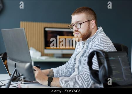Person with glasses working on laptop in modern office space with multiple screens and technological gadgets visible. Look of intense focus and determ Stock Photo