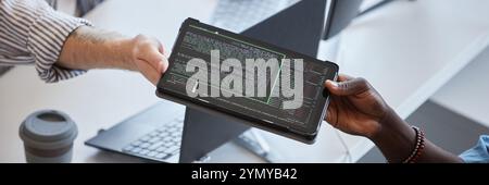 Close-up of two professionals exchanging tablet displaying code in an office environment There is emphasis on collaborative effort in a tech-related p Stock Photo