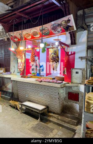 A butcher shop in Morocco is open for business, with various cuts of meat hanging from the ceiling and laid out on the counter. A cat walks by in the Stock Photo