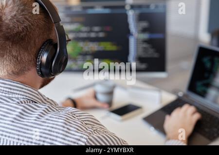 Person wearing headphones and typing on laptop while holding coffee cup, with computer screen featuring code in background. Engaging in programming in Stock Photo