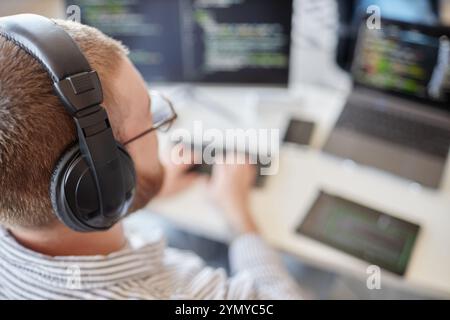Man wearing headphones, typing on keyboard and focused on coding on multiple computer screens in a well-lit workspace, engaging in software developmen Stock Photo