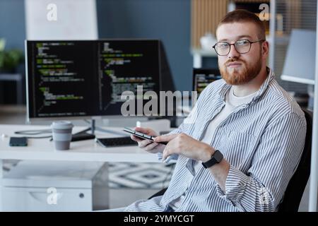 Portrait of software developer holding smartphone, sitting at desk with coding screens in background, workplace setting showing serious expression Stock Photo