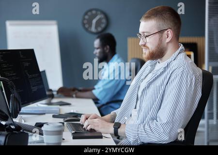 Team of software developers working on computers in modern office with focus on their tasks and coding screens visible on desks Stock Photo