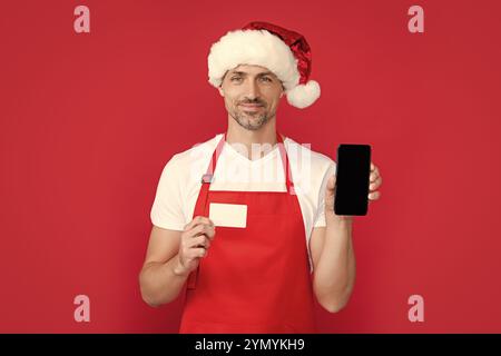 mature man in christmas santa hat and red apron hold smartphone and card. online banking Stock Photo