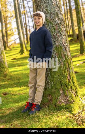 Vertical full length portrait of a caucasian happy boy with casual clothes leaning relaxed on tree trunk enjoying nature Stock Photo