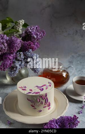 Elegant white cake with floral purple decorations on a ceramic plate surrounded by blooming lilacs, a teapot, and a cup of tea Stock Photo