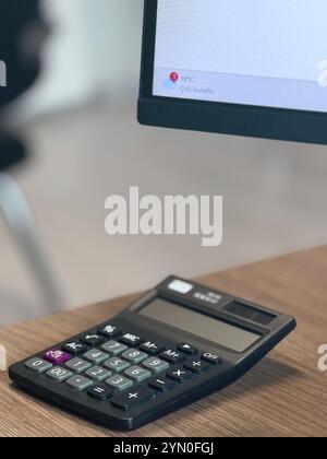 A black calculator on a desk near a computer monitor in an office environment Stock Photo