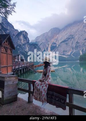 Nestled against the breathtaking mountains, a woman in a flowing dress sits peacefully at the lake edge, reflecting on nature's beauty as the soft morning light begins, Lago Di Braies Dolomites Italy Stock Photo