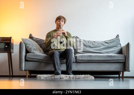 Clarinet player. Man clarinetist playing flute woodwind music instrument sitting on couch at home Stock Photo