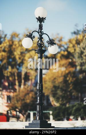 Ornate black vintage street lamp with three round frosted glass globes, standing in a park against a blurred background of autumn trees and soft blue Stock Photo
