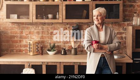 Calm senior lady standing at kitchen, drinking coffee Stock Photo