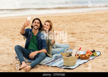 Couple enjoying a beach picnic while taking selfies during sunset Stock Photo