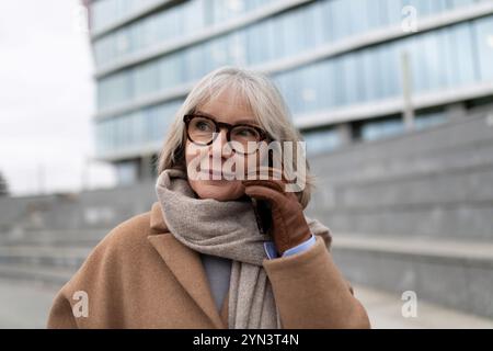 A senior woman wearing glasses talks on her smartphone while standing outside a modern building on a cool day Stock Photo