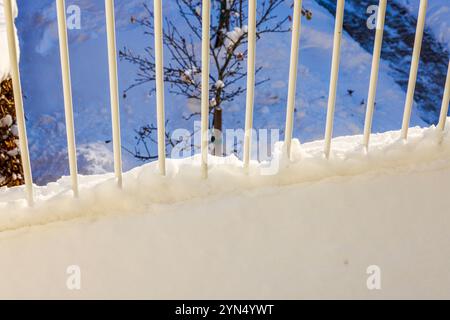Snow-covered railing on villa balcony overlooking snowy tree in garden and pathway during bright winter day. Stock Photo