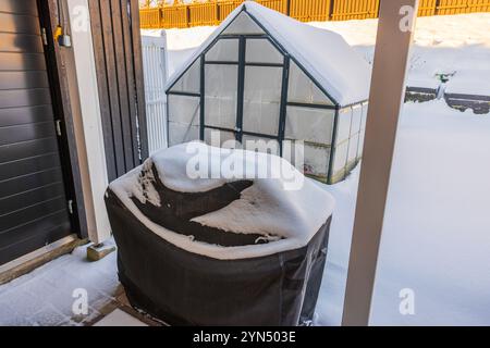 Snow-covered barbecue grill and greenhouse in backyard during cold winter morning. Stock Photo