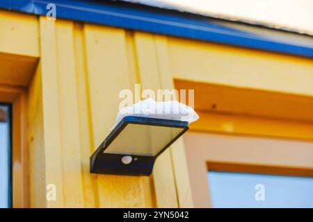 Solar outdoor LED light with motion sensor and snow resting on its surface mounted on yellow wooden wall villa. Stock Photo