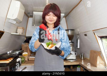 Low angle shot of Asian smiling chef adding ketchup in fresh cooked hot dog on grill in food truck kitchen Stock Photo