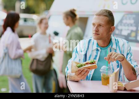 Medium close up of Caucasian young adult man relaxing while eating delicious hot dog and drinking craft lemonade Stock Photo