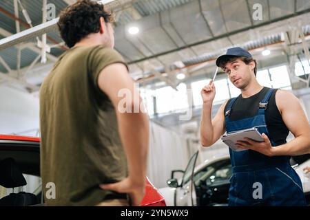 Low-angle view of male garage worker in uniform and unrecognizable man customer talking to discuss details of car repairs. Stock Photo