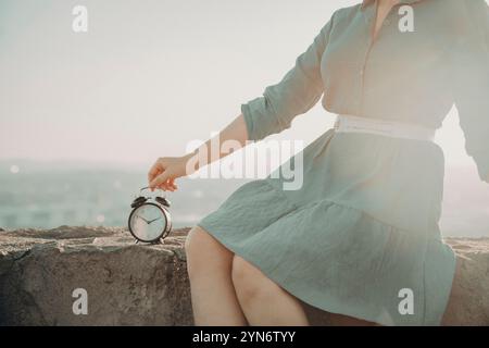 A woman is sitting on a rock with an alarm clock in her hand. The clock is set to the time of 9:00 Stock Photo