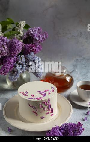 Elegant white cake with floral purple decorations on a ceramic plate surrounded by blooming lilacs, a teapot, and a cup of tea Stock Photo