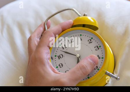 Retro alarm clock with bells on a pillow hugged by a tired man's hand. Morning routine and hard awakening concept, close-up with selective focus Stock Photo