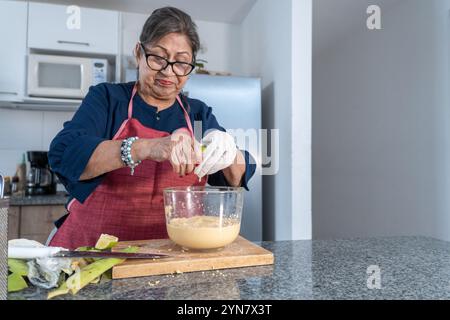 Adult woman in blue blouse and red apron squeezing a lemon into a bowl with a preparation in the kitchen with green banana peels on the counter Stock Photo