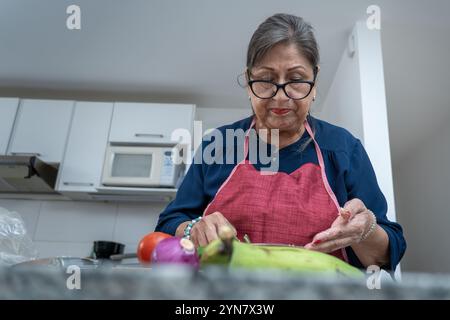 A woman is cutting up vegetables in a kitchen. She is wearing a blue apron and a red bandana Stock Photo