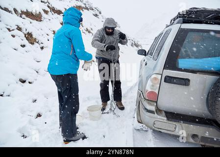 Traveling in winter, pair of friends, man and woman, putting chains on the wheels of an SUV vehicle, in order to be able to move forward over the snow Stock Photo