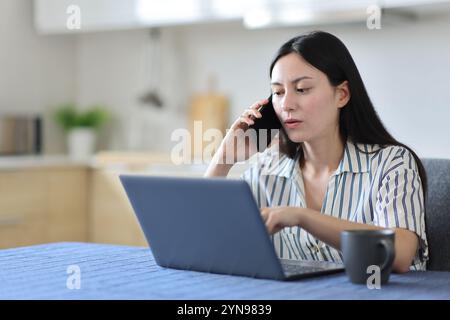 Asian woman talking on phone checking laptop solving problem in the kitchen at home Stock Photo