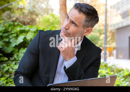 Thoughtful Businessman Outdoors Using Laptop in Urban Park Stock Photo