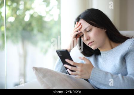 Worried asian woman checking cell phone sitting on a couch at home Stock Photo