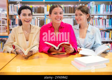 Three smiling young women reading books at library table looking at camera Stock Photo