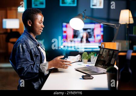 Side-view of black woman with a clipboard having a video call, discussing with group of colleagues through laptop. African american female freelancer having online meeting comparing documents. Stock Photo