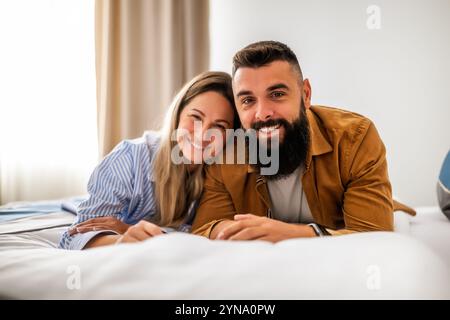 Young happy couple is lying on bed in their bedroom, looking at camera and smiling. Stock Photo